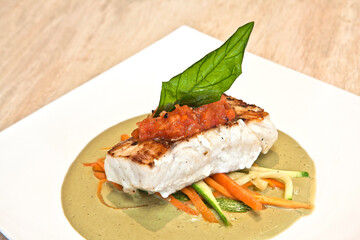 Snook fillet with epazote cream, steamed vegetables and Mexican red sauce served on a square white plate over marble background, close up view