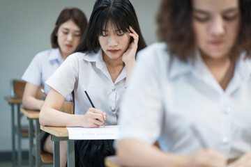 Selective focus of the teenage college students sit on lecture chairs do final examination and write on examination paper answer sheets in the classroom. University students in uniform in classroom.