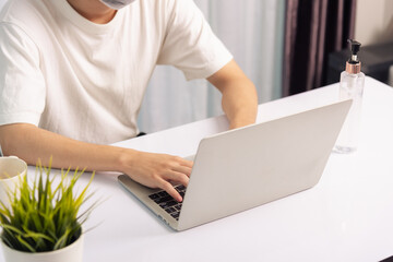 Fototapeta premium Asian business young handsome man glasses, wearing protective mask with disinfectant gel beside he work from home office remote online with laptop computer quarantines disease coronavirus or COVID-19
