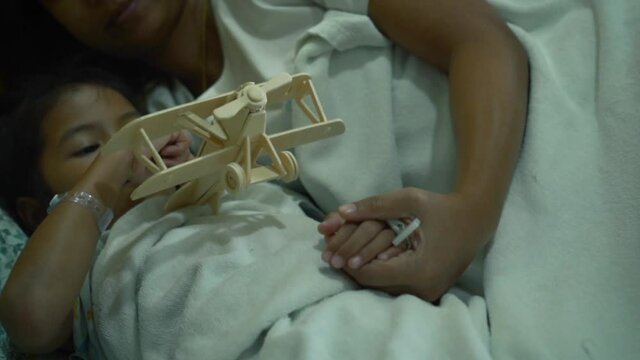 Sick asian little girl playing toy wooden airplane with her mother together while stay in the hospital bed. Therapy for patients to relax. Recovering little girl. Slow motion shot.