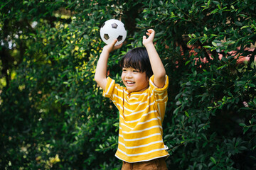 Cheerful asian kid holding football showing hands in the air at park.