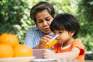 Senior elderly woman feeding orange juice to grandson at yard.