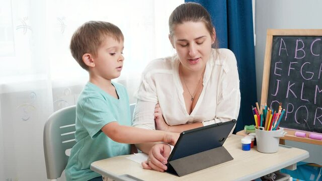 Young female teacher with little boy using tablet computer while learning alphabet at school classroom. Parenting and remote school education at home.