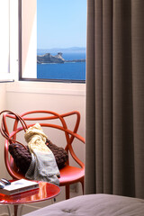 Window and curtain overlooking the sea and the cliff with red designer chair with bag and scarf on it, table with book