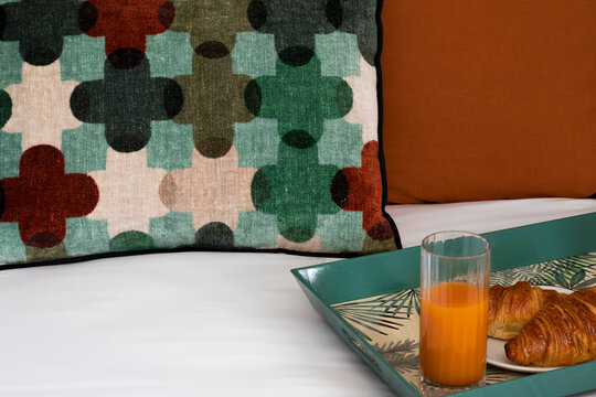 Close upl of the bed with a colorful pillow with colorful cross designs and a tray with breakfast of croissants and orange juice