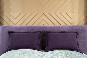 Close up view of purple pillows and headboard of an empty double bed with gray bedspread. Designer wall with volumetric beige stucco molding in the form of rhombuses.