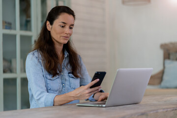 Thinking woman with phone and computer sitting on the table. Working at home. Home blurred background.