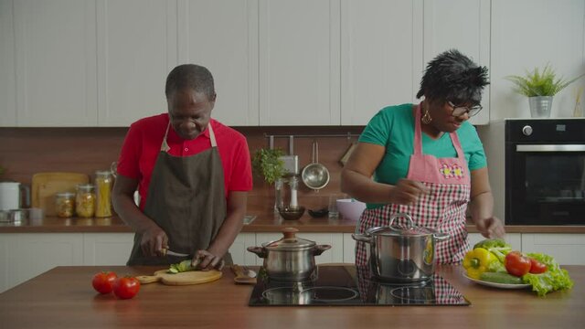 Concentrated senior african male wearing apron, chopping cucumber on cutting board with busy attractive elderly wife cooking meal while retired couple preparing dinner together in domestic kitchen.