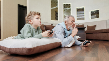 Obraz premium Lazy weekend with grandpa. Grandfather and his cute little grandson laying together on the floor and playing video games, spending time at home