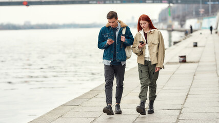 Full length shot of two teenagers totally absorbed in their smartphones, ignoring each other while walking along the riverside together
