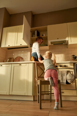 Full length shot of two naughty kids, brother and sister trying to find something sweet in the kitchen cupboard. Little girl helping her brother by holding his hand