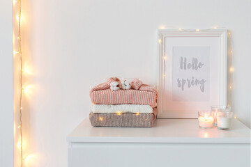 Frame with text HELLO SPRING, stack of knitted sweaters in pastel colors, candles, glowing garland...