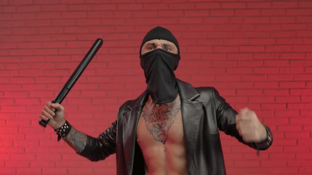 a male criminal wearing tattoos a leather raincoat and a balaclava with a baton and a weapon