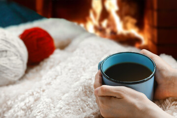 Close up of the young woman hands sitting at the winter warm blanket with a cup of coffee and skeins of yarn. Fireplace on background. 