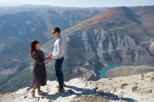 Lovely couple joins hands standing on edge of rocky cliff against mountainous landscape with small blue river in canyon at bright sunlight