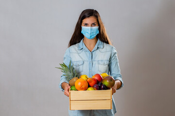 Female with surgical mask holding wooden box full of fresh fruit with copy space. Positive brunette with woody crate filled with heatlhy sweet food during pandemic. Brown hair lady with respiratory