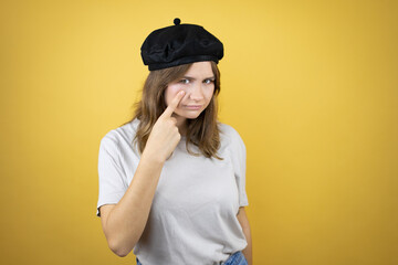 Beautiful young caucasian girl wearing french look with beret over isolated yellow background Pointing to the eye watching you gesture, suspicious expression