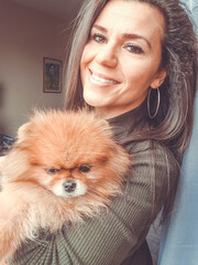 Smiling  beautiful woman hugging  cute fluffy  pomeranian spitz dog wrapped in white blanket  looking  out the window on a   cold winter day.  