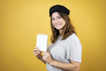 Beautiful young caucasian girl wearing french look with beret over isolated yellow background smiling and showing blank notebook in her hand