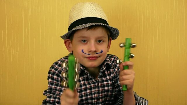 A funny Caucasian preschooler boy in an elegant hat with painted mustache holds a jingle stick in his hands. The child plays with bells and sticks. Preparation for the carnival. Selective focus