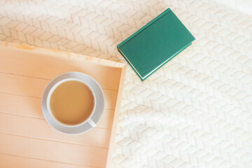 Wooden tray, a cup of coffee with milk and a green book on a light blanket. The concept of morning, home comfort. Top view, minimalism, copy space, flat lay.