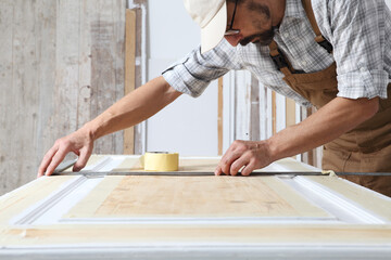 Male carpenter working the wood in carpentry workshop, taking measures on wooden door with a metal...