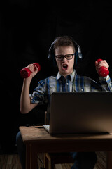 male young student with two dumbbells in front of laptop at home schooling