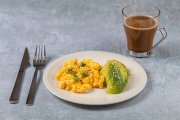 Keto diet breakfast scrambled eggs and avocado with olive oil and a cup of coffee over a clear grey background.