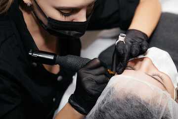 Fototapeta na wymiar Professional beautician master of permanent makeup in black medical mask and gloves makes tattooing eyebrows. Concept of beauty salon and permanent make-up. New reality in beauty world. Covid 19