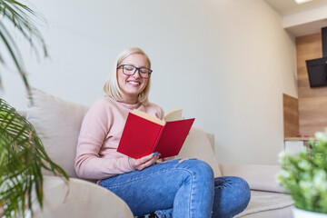 Attractive woman sitting on bed in the morning, drinking tea, reading book, casual style, blue jeans, pink sweater, feeling comfortable at home, having rest, smiling