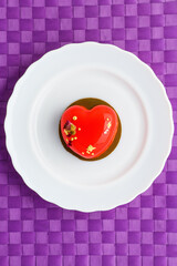 A cake in heart shape. One heart-shaped mousse cupcake on a plate on a purple background. Valentine's Day. Vertical photo