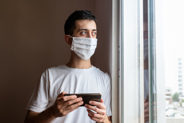 Young man in quarantine holding his smartphone and looking outside through the window