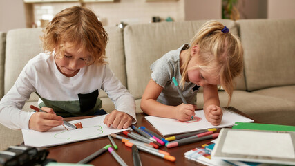 Cute little children, boy and girl looking focused while drawing on paper using marker pen, sitting together on a couch at home