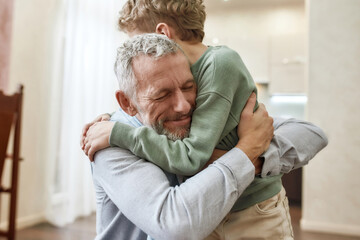 Emotional meeting. Happy grandfather embracing his cute little preschool grandson while standing together at home - obrazy, fototapety, plakaty