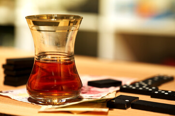 Tea glass with black tea stands next to a domino on a pile of Turkish paper money on a grained wooden board, blurred background
