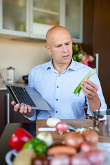 Brutal man in kitchen preparing food and uses laptop.