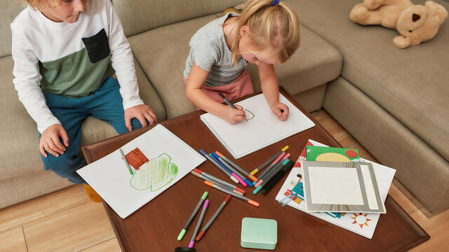 Cropped shot of little kids, boy and girl drawing on paper using marker pen, sitting together on a sofa at home