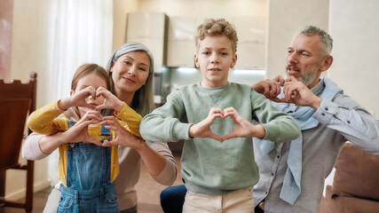 Happy caucasian family, grandparents and grandchildren making heart sign with hands and smiling at camera while sitting on the floor at home