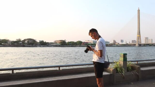 The young man takes pictures on camera on park embankment in Bangkok near Chao Phraya River 