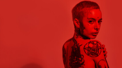Tattoo and piercing. A white woman with tattoos and piercing standing topless with her back to a camera looking into it with her hands covering breasts