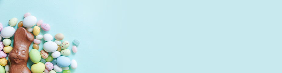 Happy Easter concept. Preparation for holiday. Easter candy chocolate eggs bunny and jellybean sweets isolated on trendy pastel blue background. Simple minimalism flat lay top view copy space. Banner