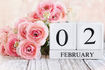 White wood calendar blocks with the date February 2nd and pink ranunculus flowers over a wooden table. Selective focus with blurred background. 