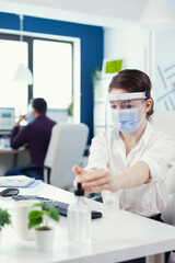Office worker following safety precaution during global pandemic with coronavirus applying sanitizer. Businesswoman in new normal workplace disinfecting while colleagues working in background.