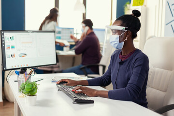 African woman typing on computer at workplace wearing face mask as safety precaution agasinst covid19. Multiethnic team in new normal business financial office checking reports, analysing datas