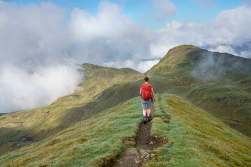 Fototapeta premium A female hiker walking over the narrow mountain ridge of the summit of Meall Garbh towards Beinn Nan Eachan near Loch Tay in the Scottish Highlands, UK mountains.