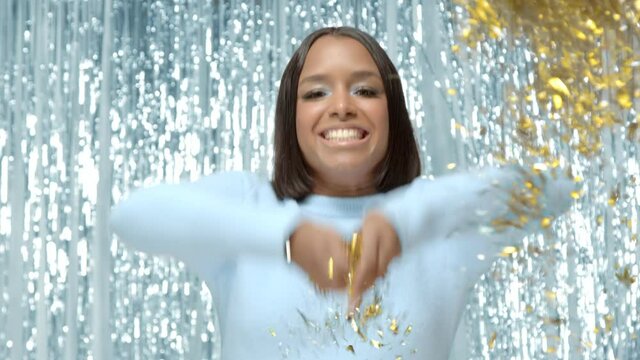Joyful festive portrait of mixed race black woman laughing and scattering golden sparkles. Blue sweater and light blue shimmery background. Middle still shot 4k high quality video footage.