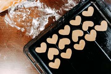 Cookies in shape of heart for the Saint Valentine's Day. Dough, flour, baking pan, round wooden cutting board and rolling pin on the table.