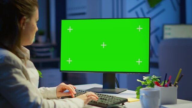 Freelancer working in front of green screen display sitting at desk in business office late at night. Businesswoman watching desktop laptop monitor with green mockup, chroma key, working overtime.