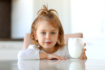 Cute little girl with glass of milk at table in kitchen. White home concept