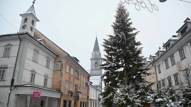Snow falling in small historic town Kranj, Slovenia. Snowstorm in winter season. December Christmas decoration with tall spruce. Old medieval town in Europe. Tilt down, wide angle
